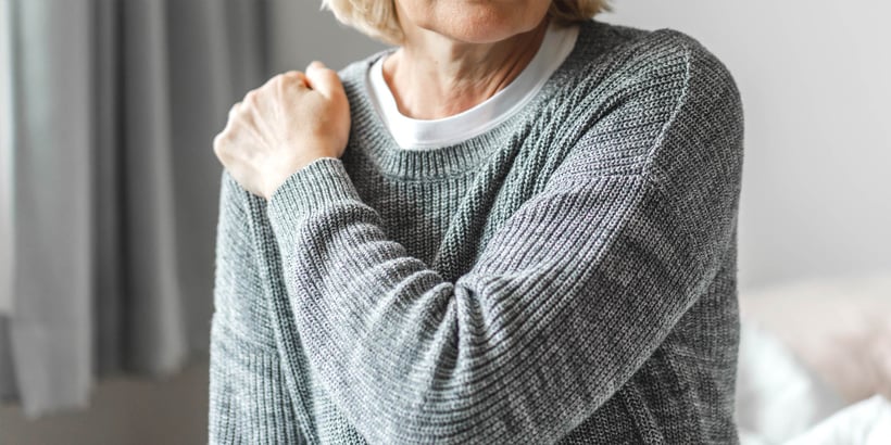 Eine ältere Frau im grauen Pulli hält ihre rechte Schulter. 